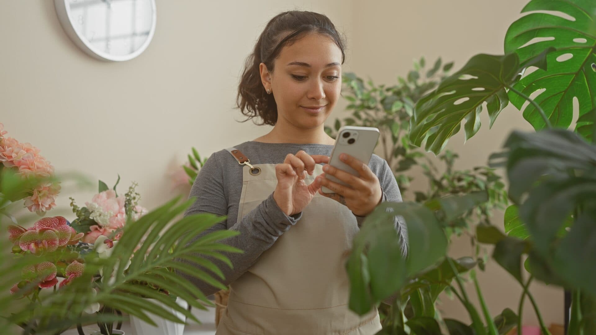 Joven latina con mandil revisa mensajes en su celular rodeada de plantas en su tienda