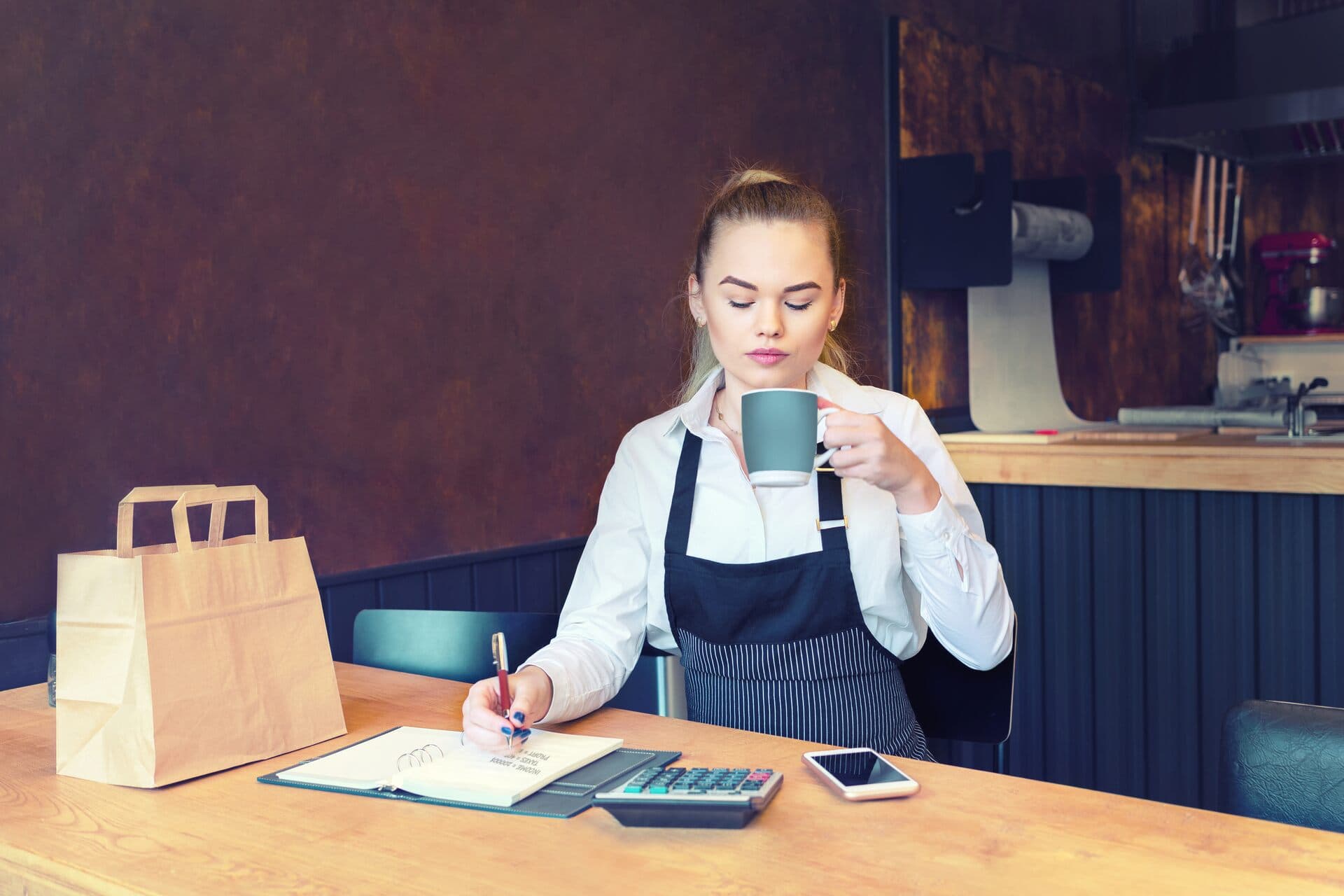Dueña de una cafetería toma café y anota gastos en su libreta con calculadora y celular