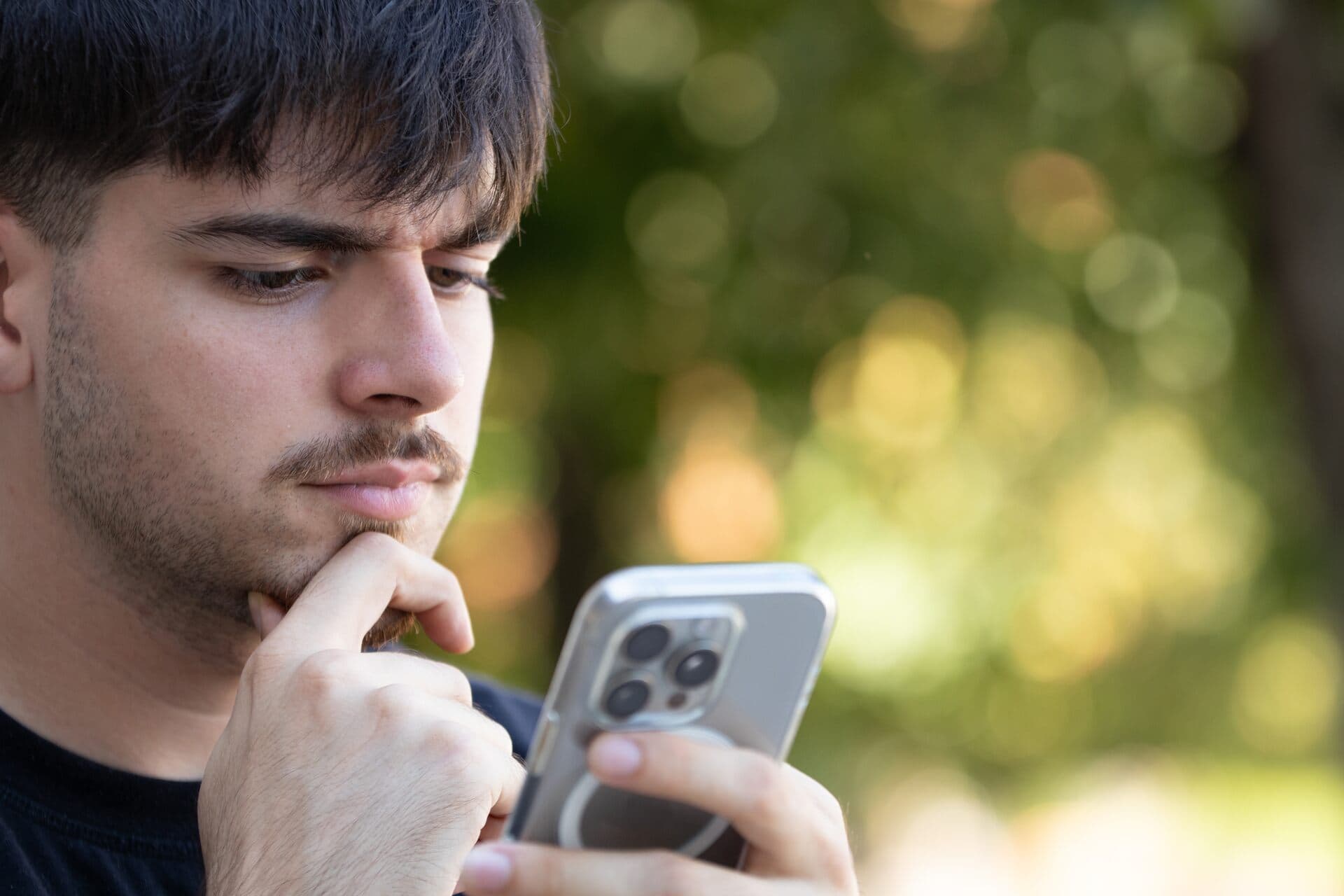 Joven con la mano en la barbilla mira impaciente la pantalla de su celular al aire libre