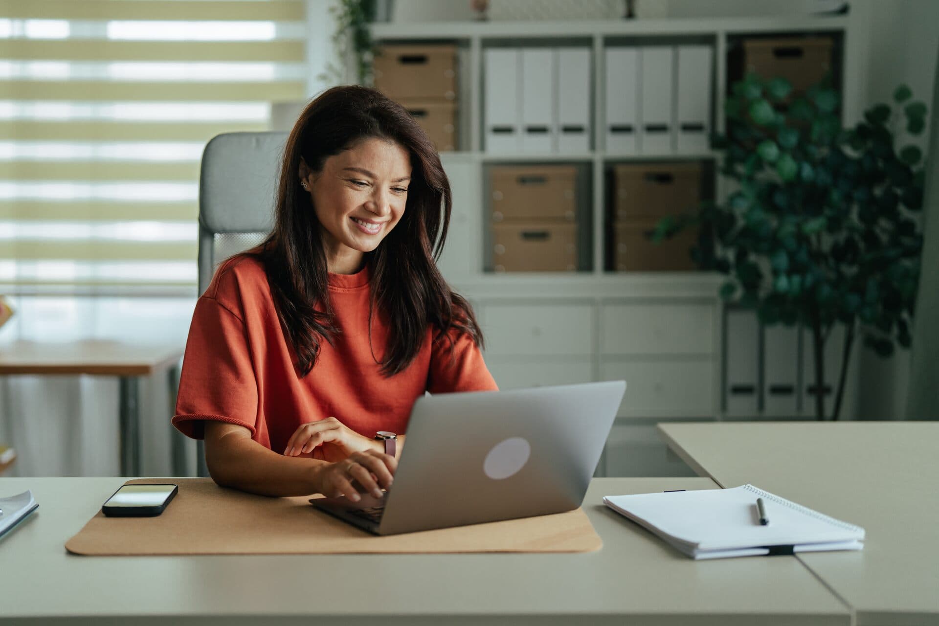 Mujer sonriente trabaja en su laptop en una oficina luminosa con cajas de archivo y plantas