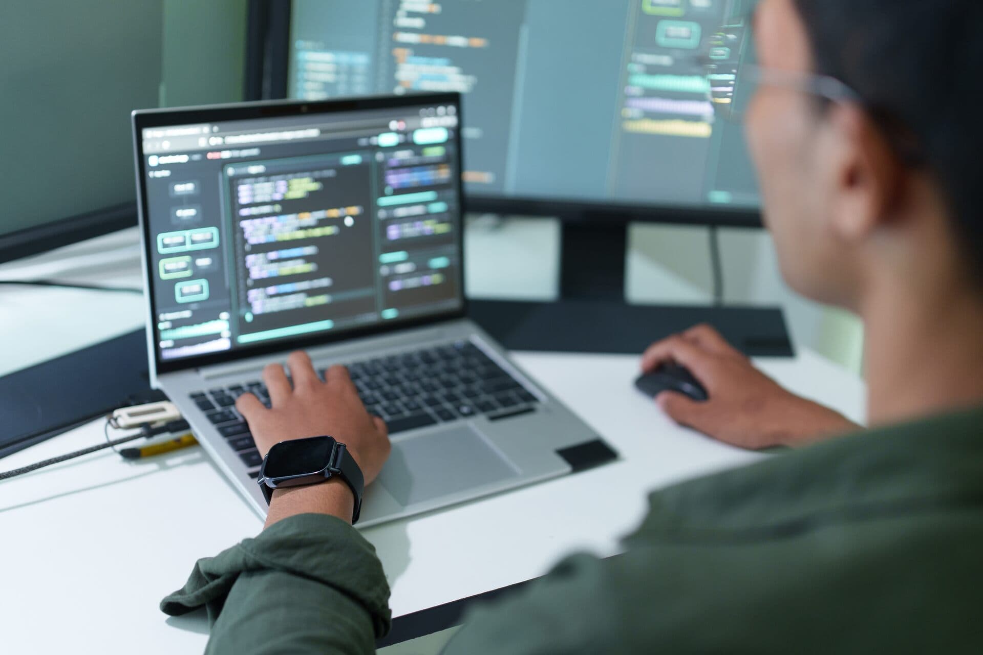 Vista sobre el hombro de un programador que escribe código en una laptop junto a otro monitor