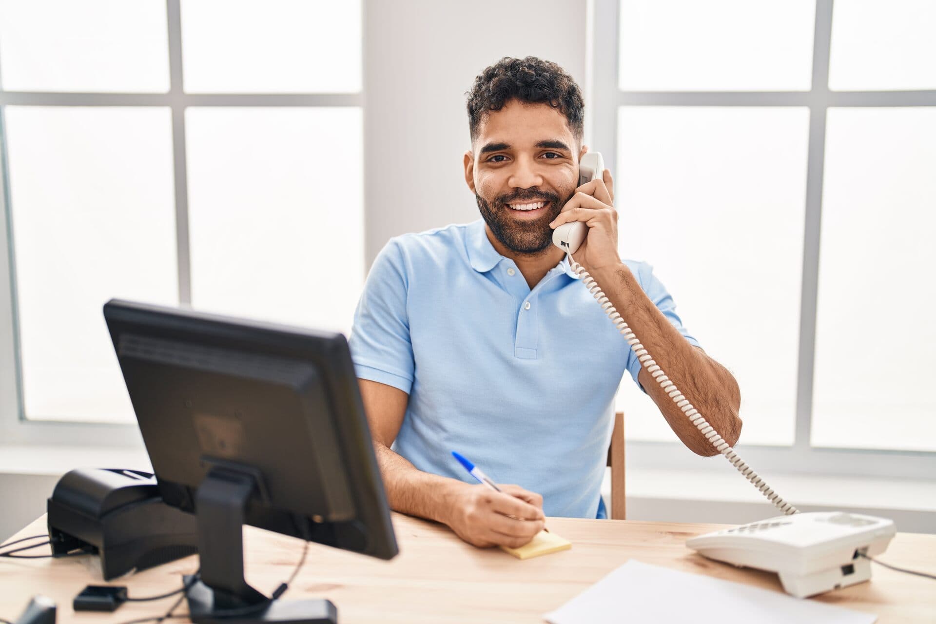 Hombre latino sonriente atiende una llamada por teléfono fijo en su escritorio de oficina