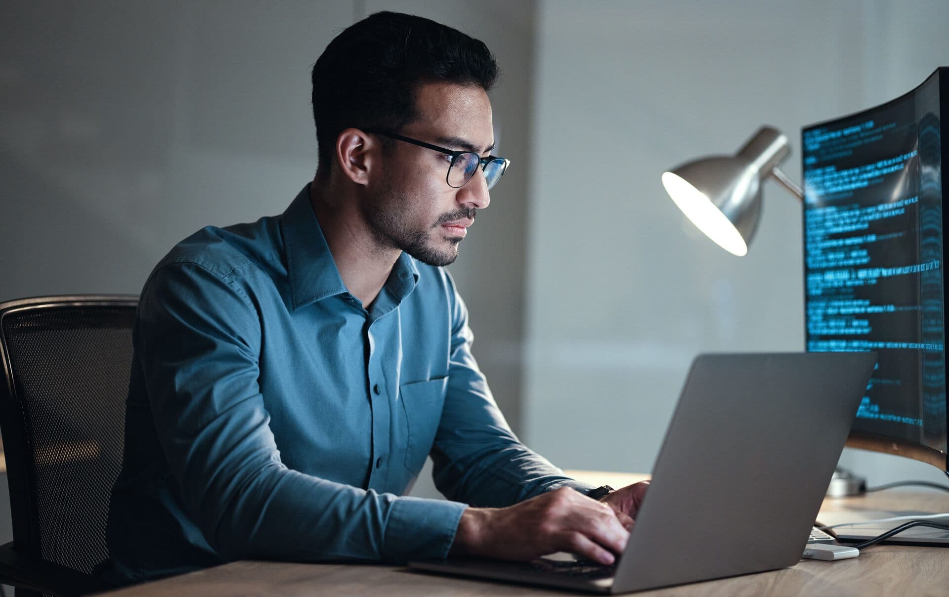 Desarrollador concentrado trabaja de noche en su laptop junto a un monitor con código