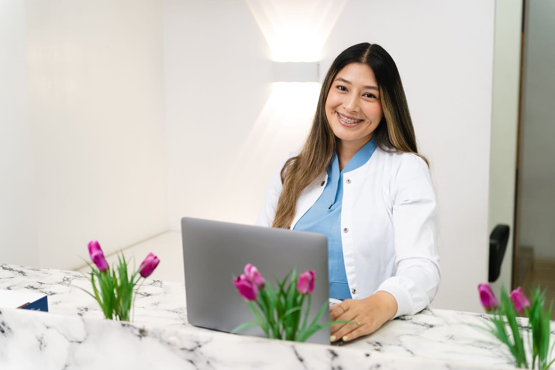Smiling receptionist behind a clinic front desk with a laptop and tulips