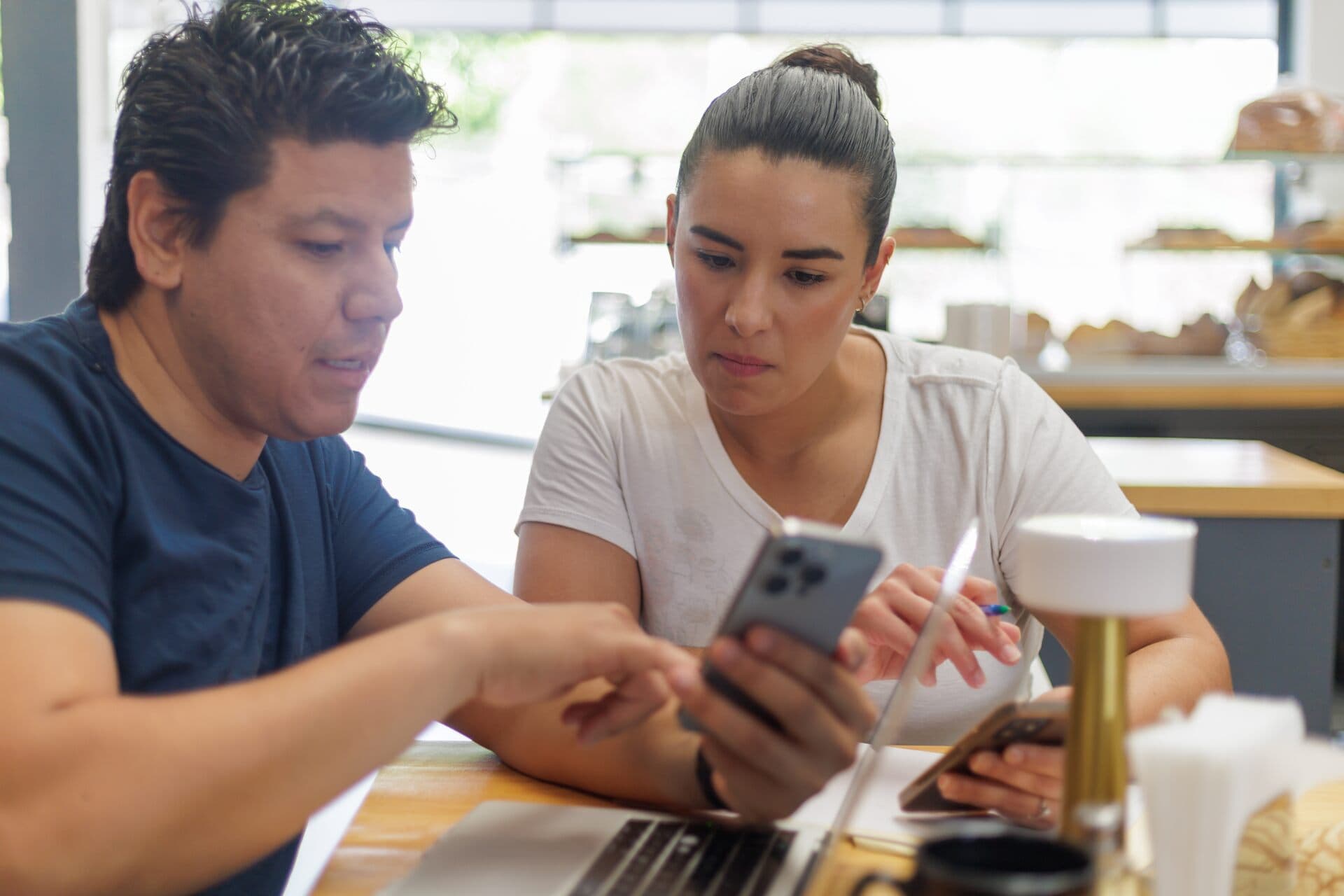 Pareja latina revisa ideas en el celular y la laptop mientras toma notas en una cafetería