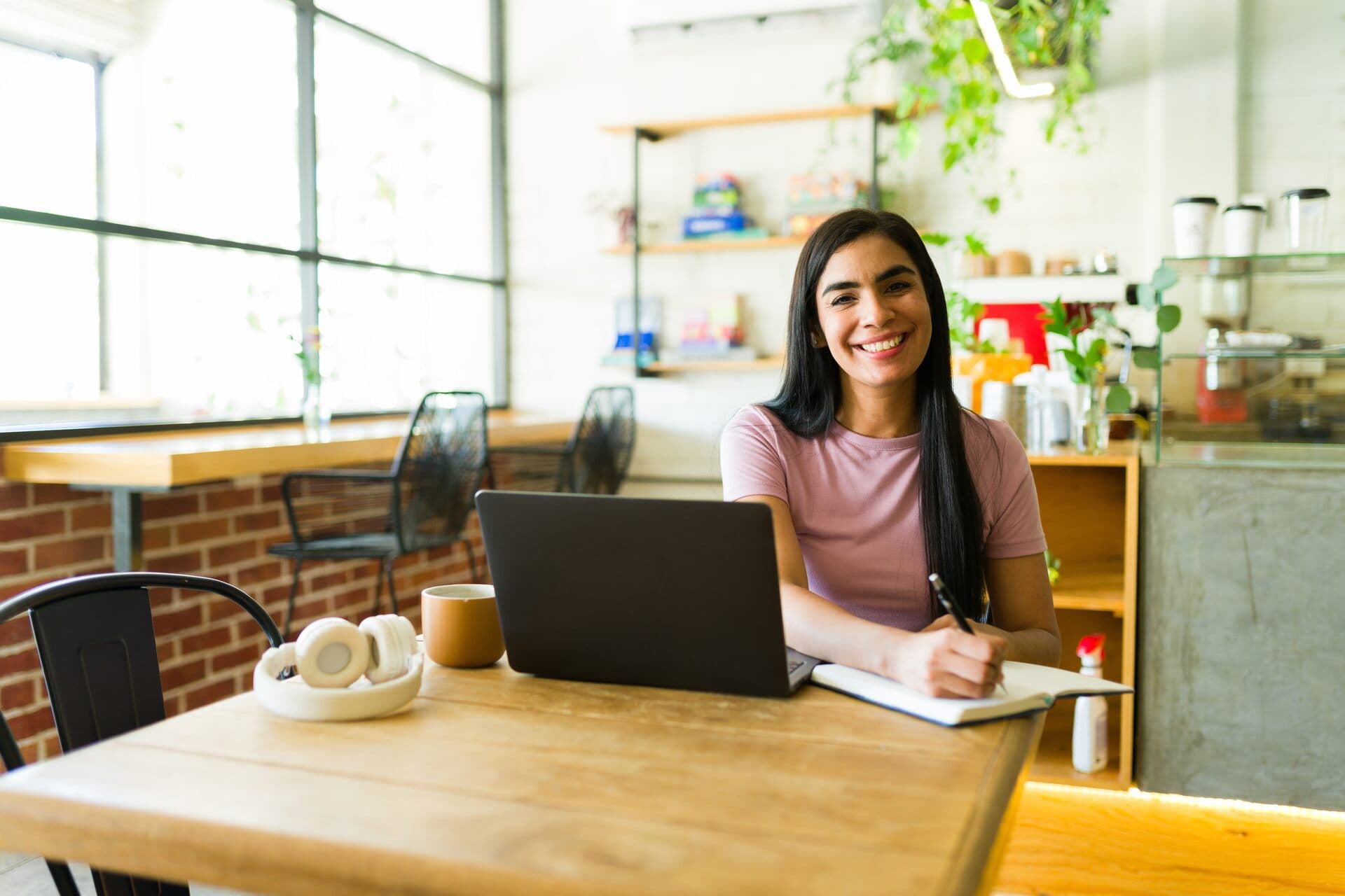 Emprendedora latina sonriente trabaja con su laptop y toma notas en un espacio luminoso
